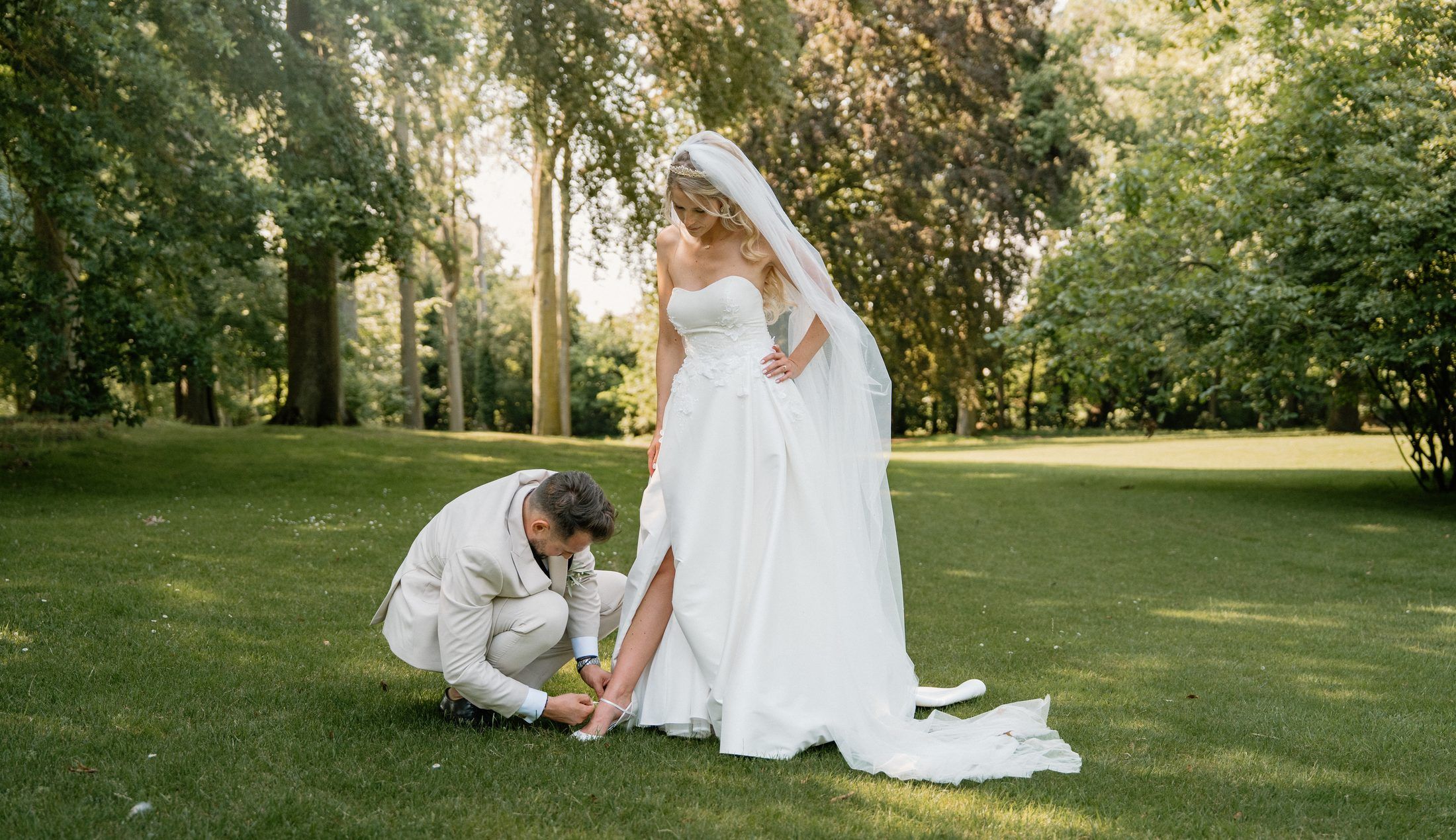 Le marié agenouillé ajuste la chaussure de la mariée sur la pelouse, arbres alentour