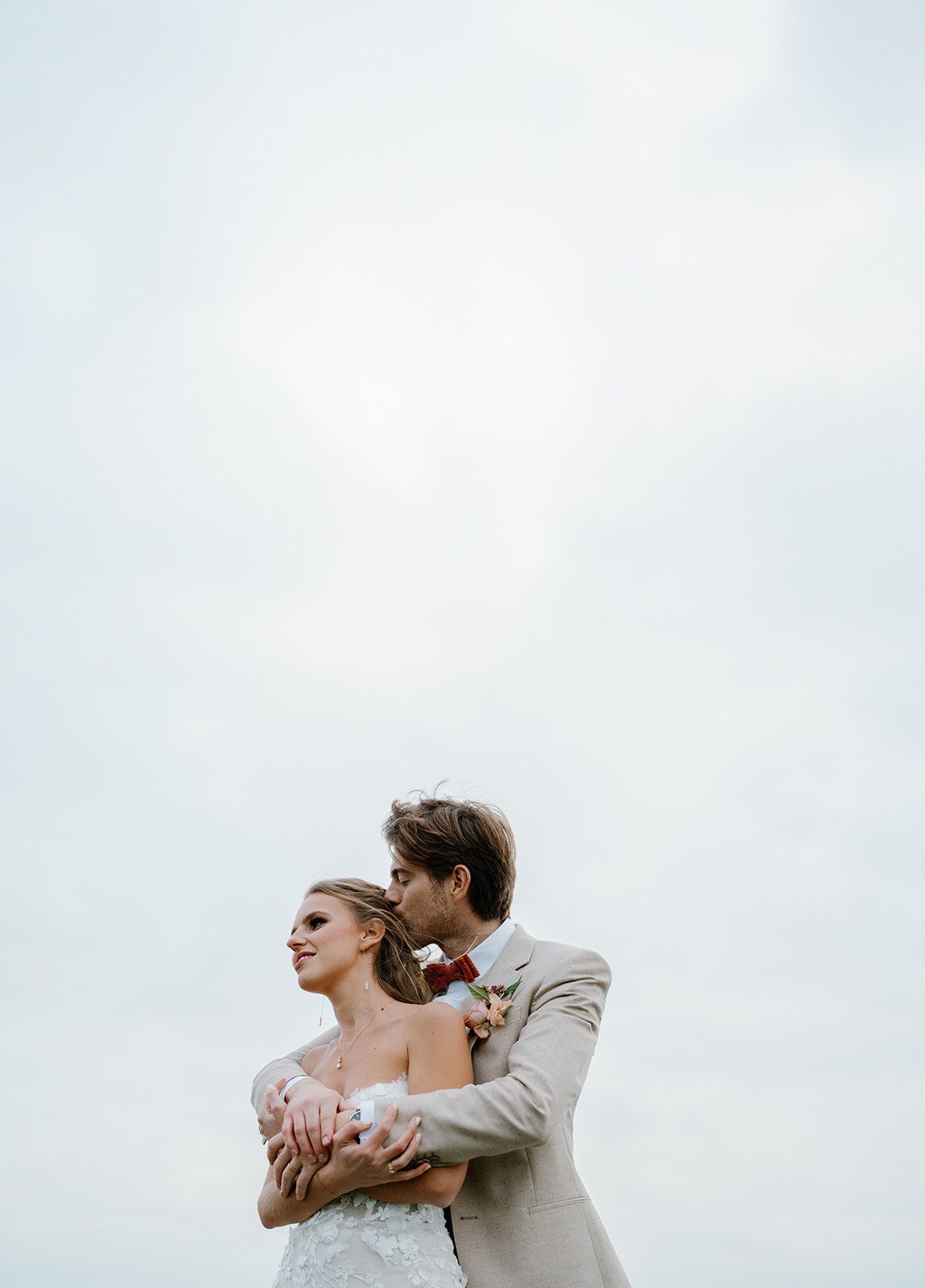 Le marié pose un baiser sur la tempe de la mariée, ciel dégagé en arrière-plan